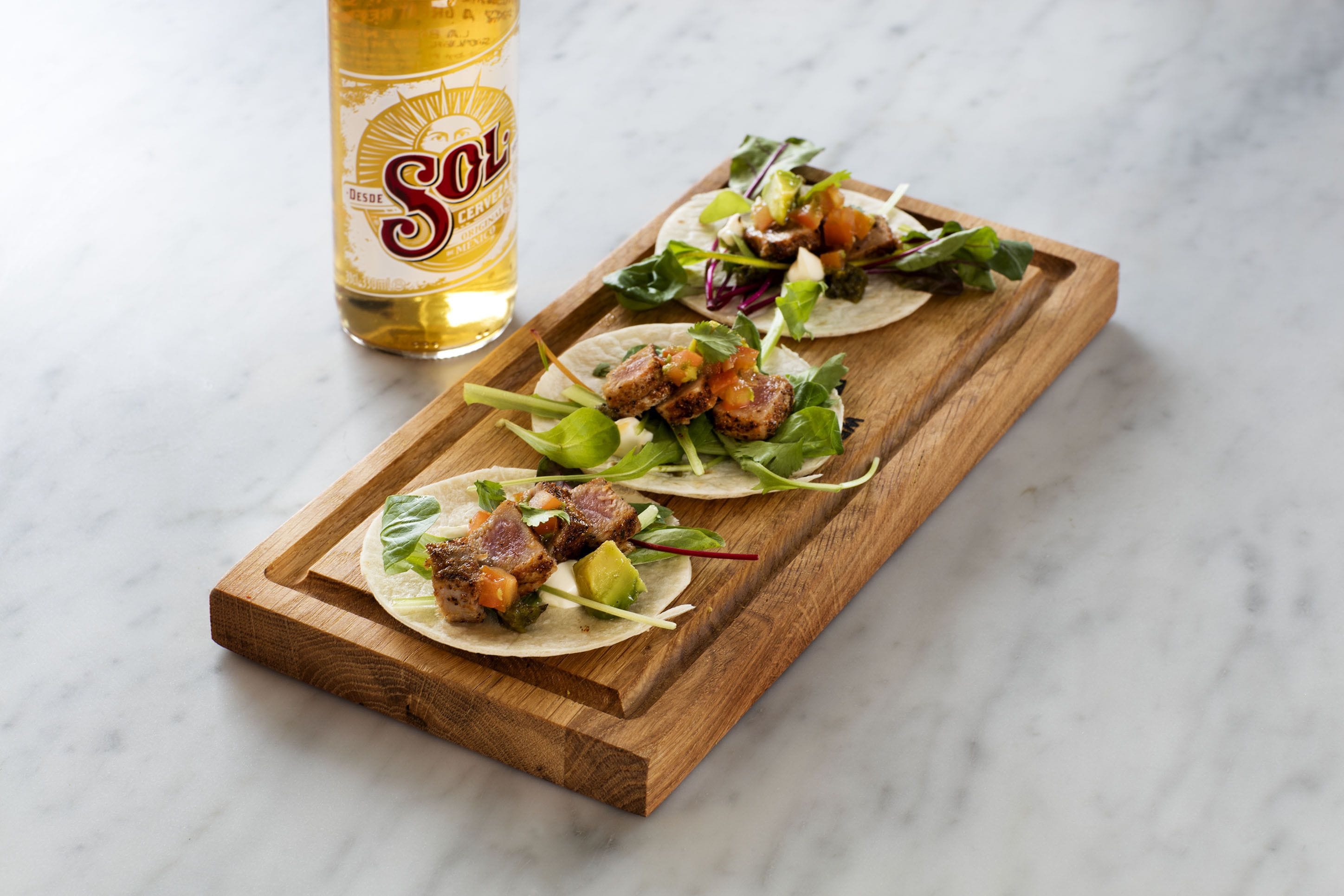 Een houten dienblad met drie taco's en een fles bier.