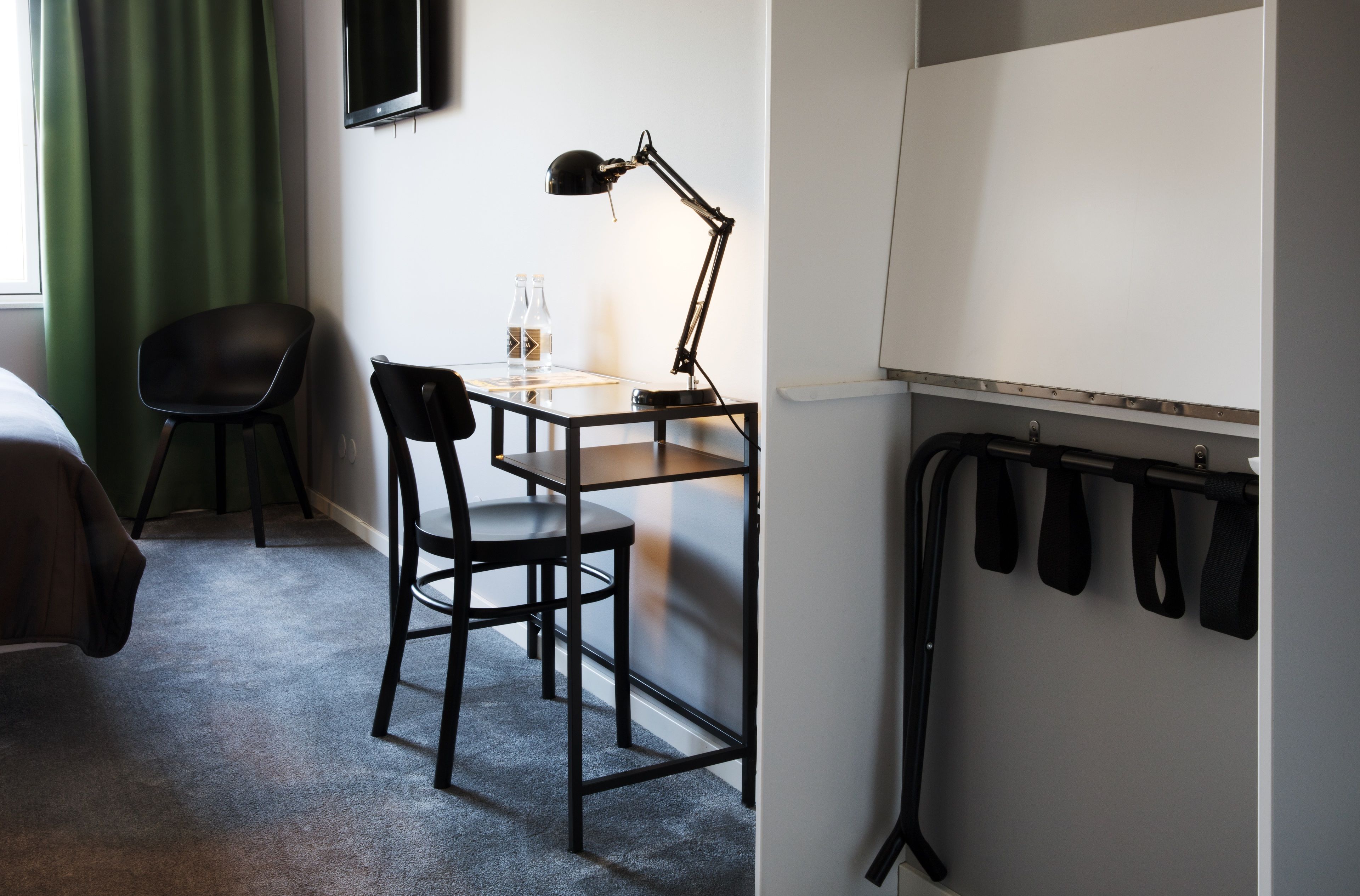 Quarto de hotel com escrivaninha, duas cadeiras, televisão fixada na parede e um cabideiro com vários penduradores.