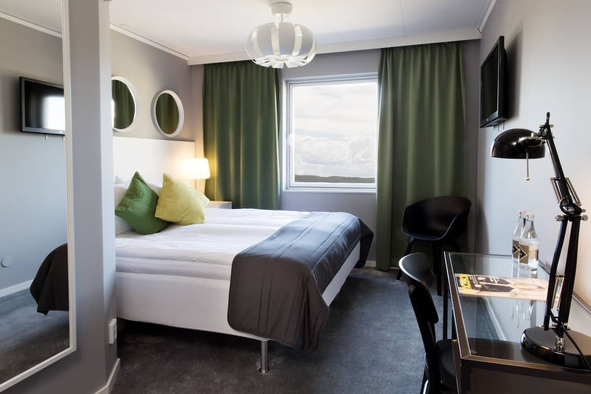 Habitación de hotel con cama, escritorio con lámpara, silla, televisor y ventana con cortinas.