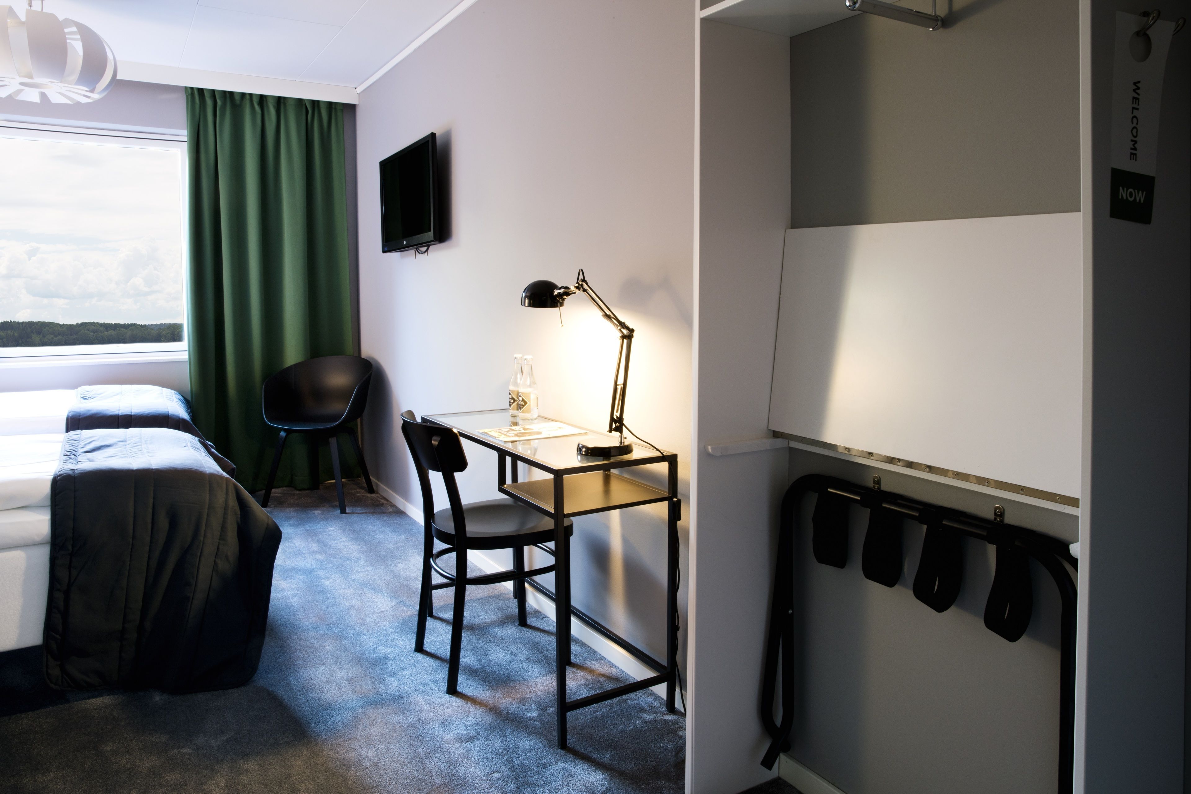 Una habitación de hotel con una cama, un escritorio con lámpara, una silla, un televisor y un perchero.