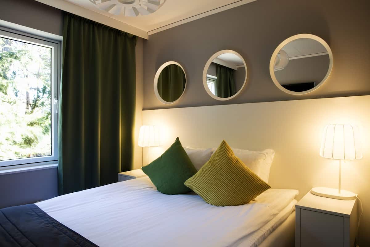 Una cama bien hecha con almohadas verdes y amarillas, una lámpara de noche y tres espejos redondos en la pared.