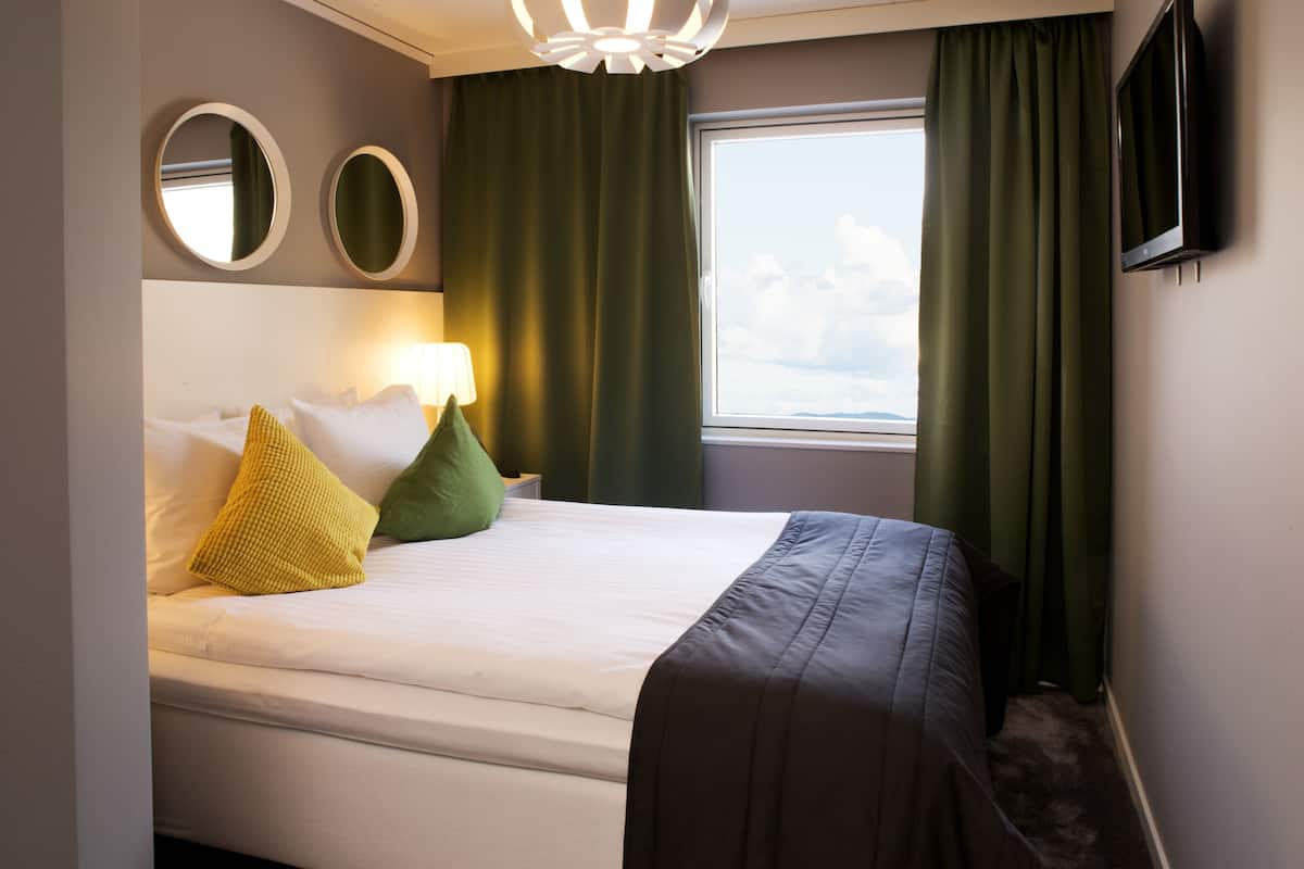Habitación de hotel con cama, dos mesitas de noche, lámpara, televisor y ventana con cortinas.