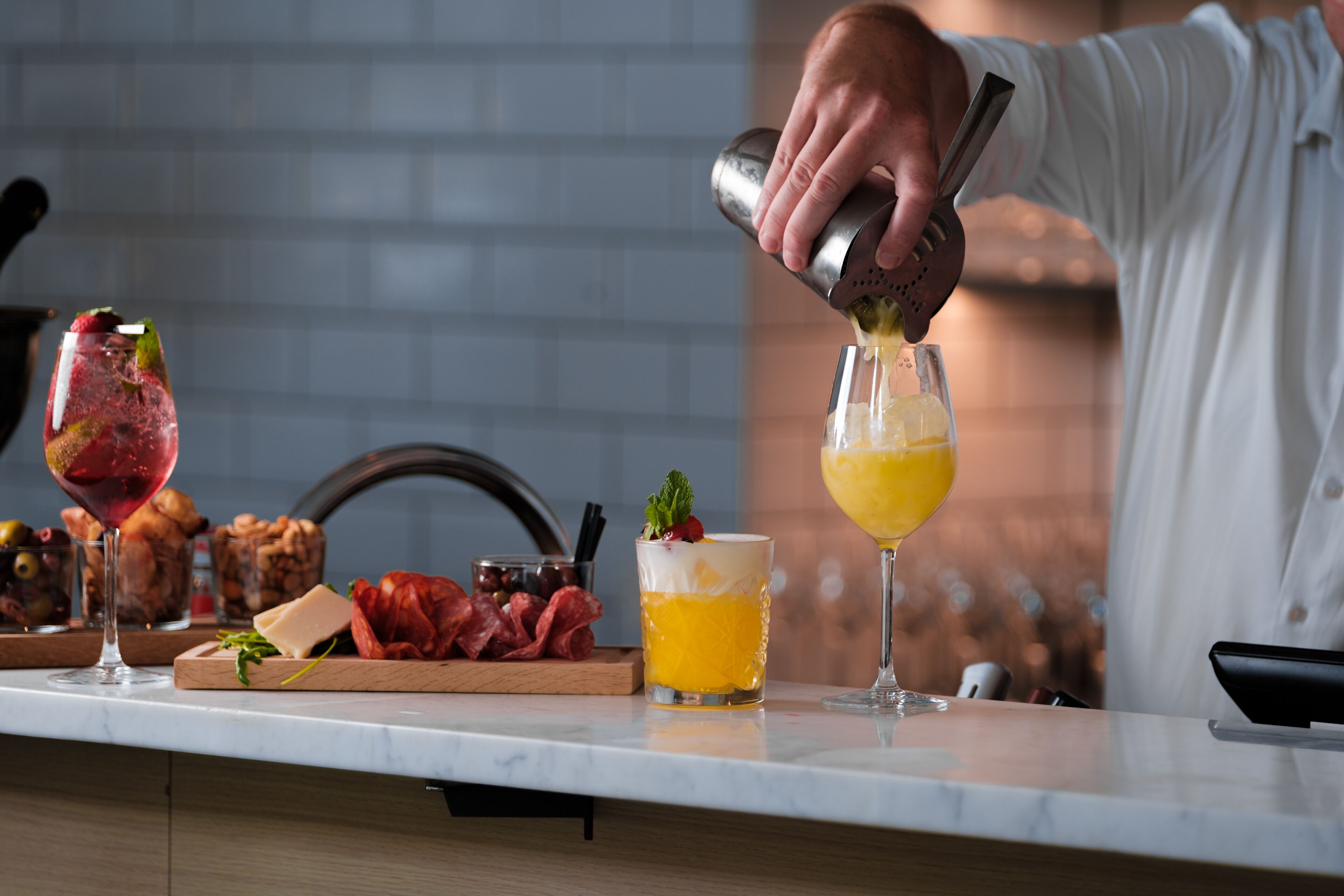 A bar setting with a bartender preparing drinks. There are two glasses on the counter, one with a red drink and another with a yellow drink. A wooden board with various food items is also visible.