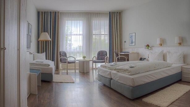 A modern hotel room with a large bed, a desk, two chairs, a small table, and a window with curtains.