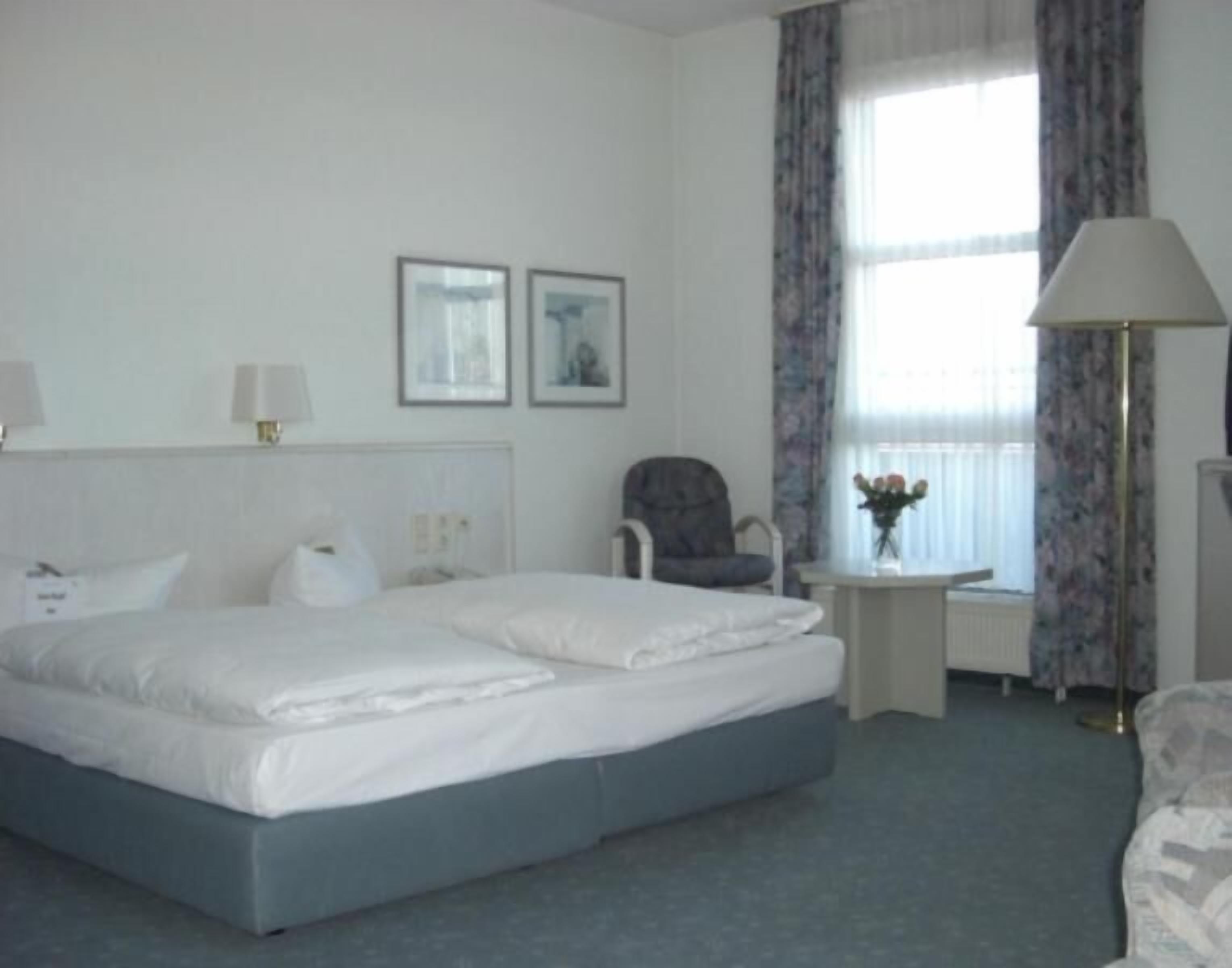 Habitación de hotel con una cama grande, una silla, una mesita con un jarrón de flores y dos cuadros en la pared.
