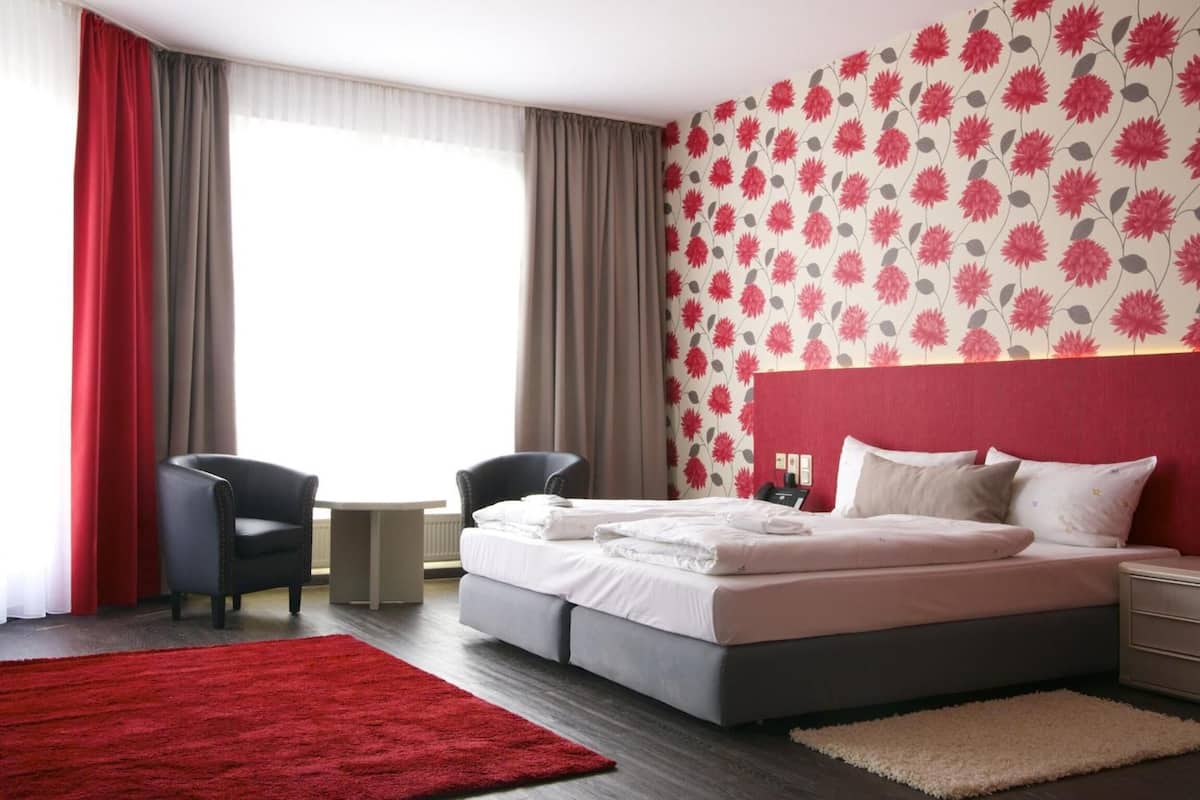 A hotel room with a bed, two chairs, a small table, and a red and white floral wallpaper.