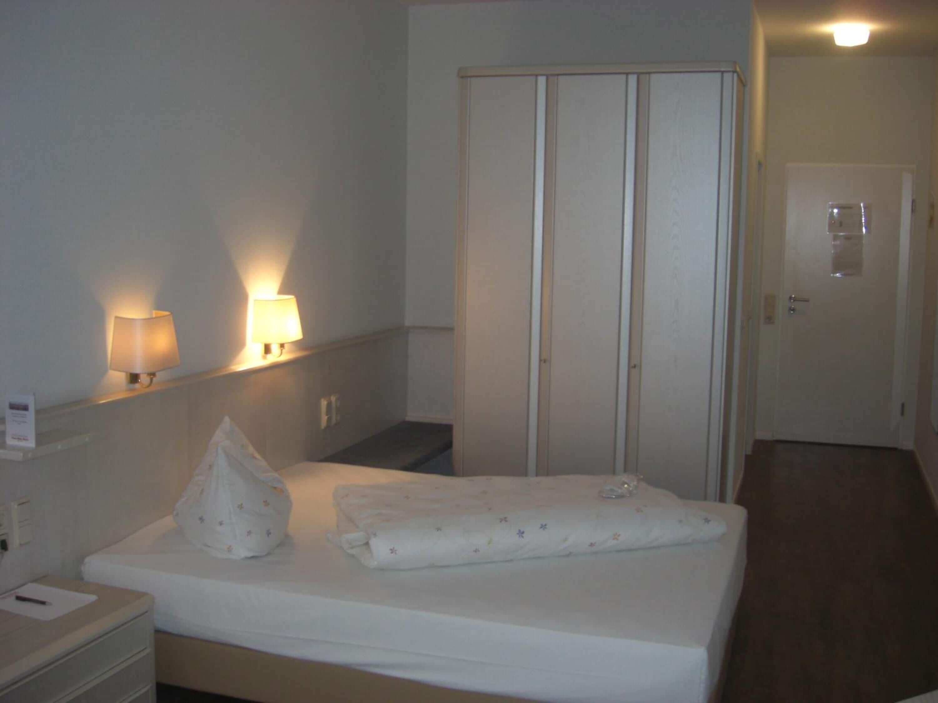 Una habitación de hotel con cama, lámparas de noche, un armario y una puerta.