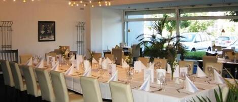 Un restaurante con largas mesas dispuestas para comer, con manteles blancos, servilletas doradas y plantas en macetas.
