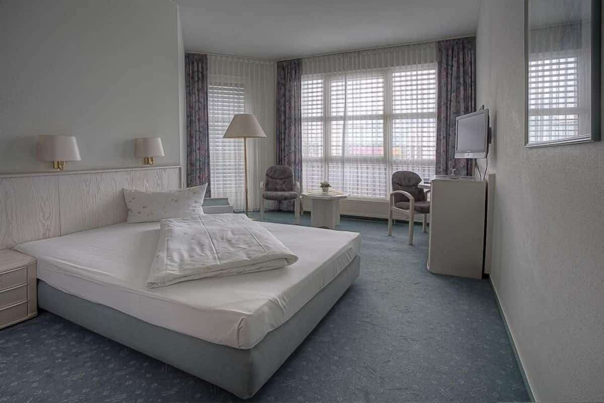 A hotel room with a large bed, two bedside lamps, a desk with a chair, a television, and large windows with curtains.