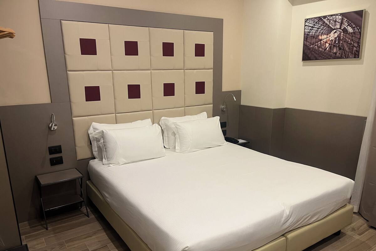A hotel room with a large bed, a headboard with square panels, a bedside table, and a wall-mounted artwork.
