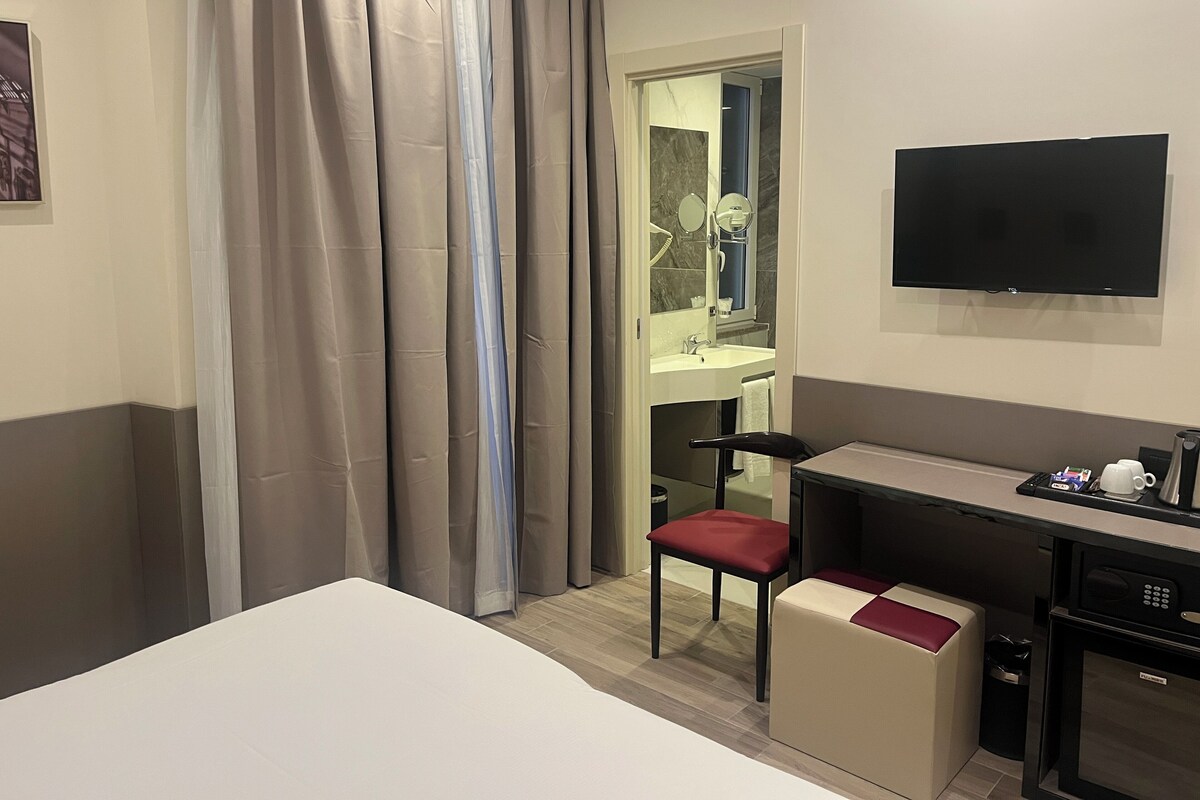 Quarto de hotel com cama, escrivaninha com cadeira, televisão e pequeno espaço de cozinha.