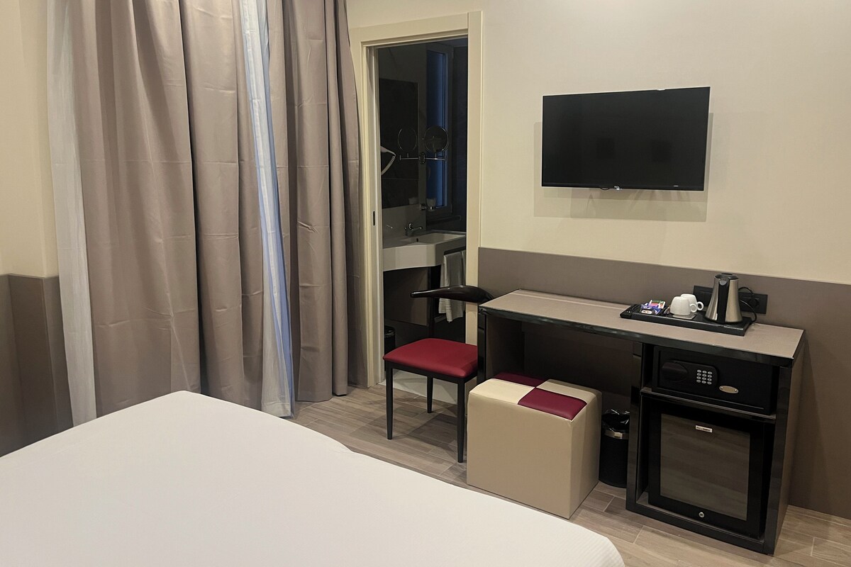 Quarto de hotel com cama, escrivaninha, cadeira, televisão e minibar.
