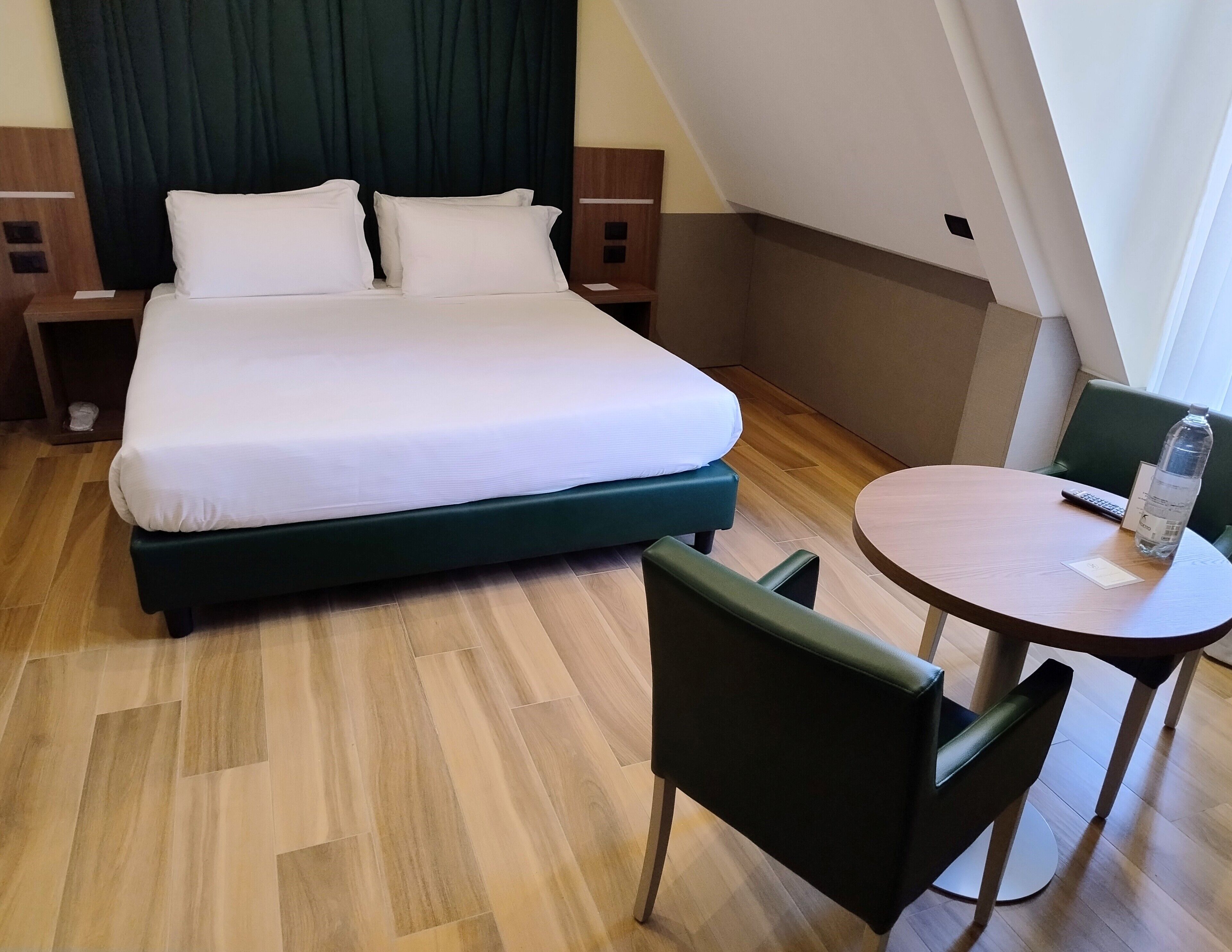 Una camera d'albergo con un letto, un tavolino con sedie, una bottiglia d'acqua e un telecomando.