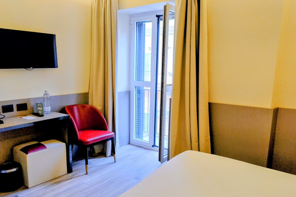 Quarto de hotel com cama, escrivaninha, cadeira, televisão e janela com cortinas.