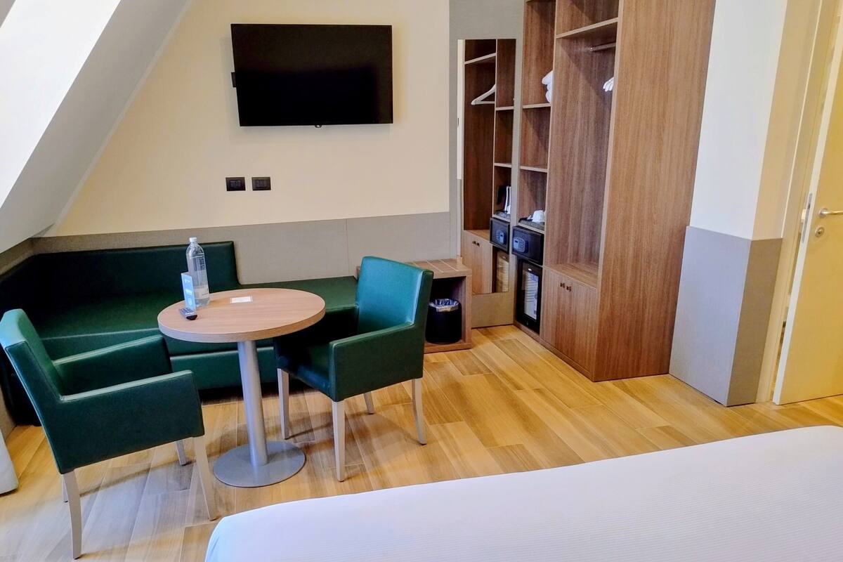 A modern hotel room with a lofted ceiling, a flat-screen TV mounted on the wall, a dining area with a round table and two chairs, a wardrobe with hanging space, and a bed with a white mattress.