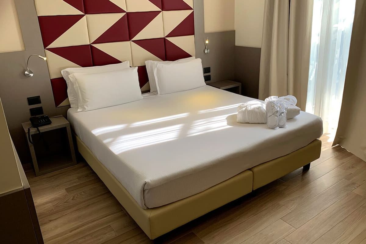 A hotel room with a large bed, a geometric-patterned headboard, bedside lamps, and a view of the outdoors.