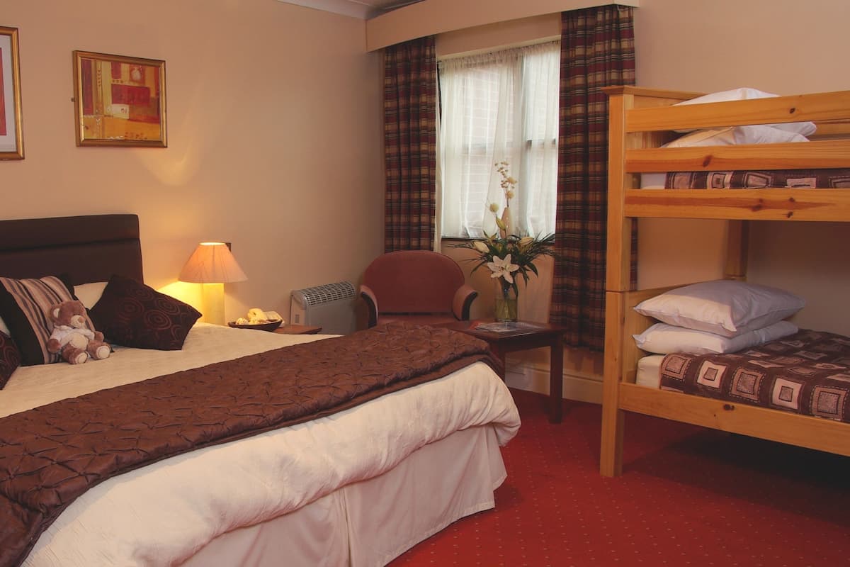 Quarto de hotel com uma cama grande, uma cama menor à direita, uma cadeira, uma escrivaninha com uma luminária e uma janela com cortinas.