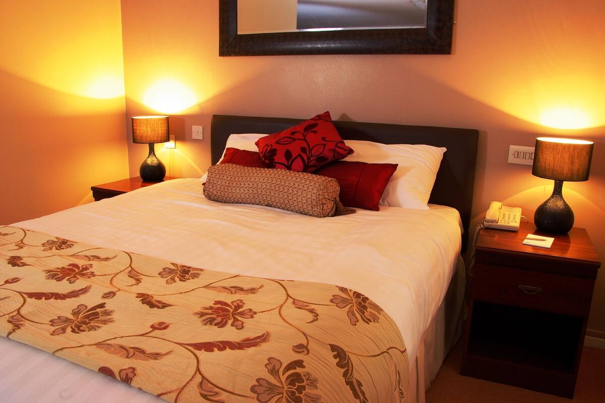 Cama bem arrumada com roupa de cama estampada com flores, duas luminárias de cabeceira, um telefone e uma gaveta de criado-mudo.
