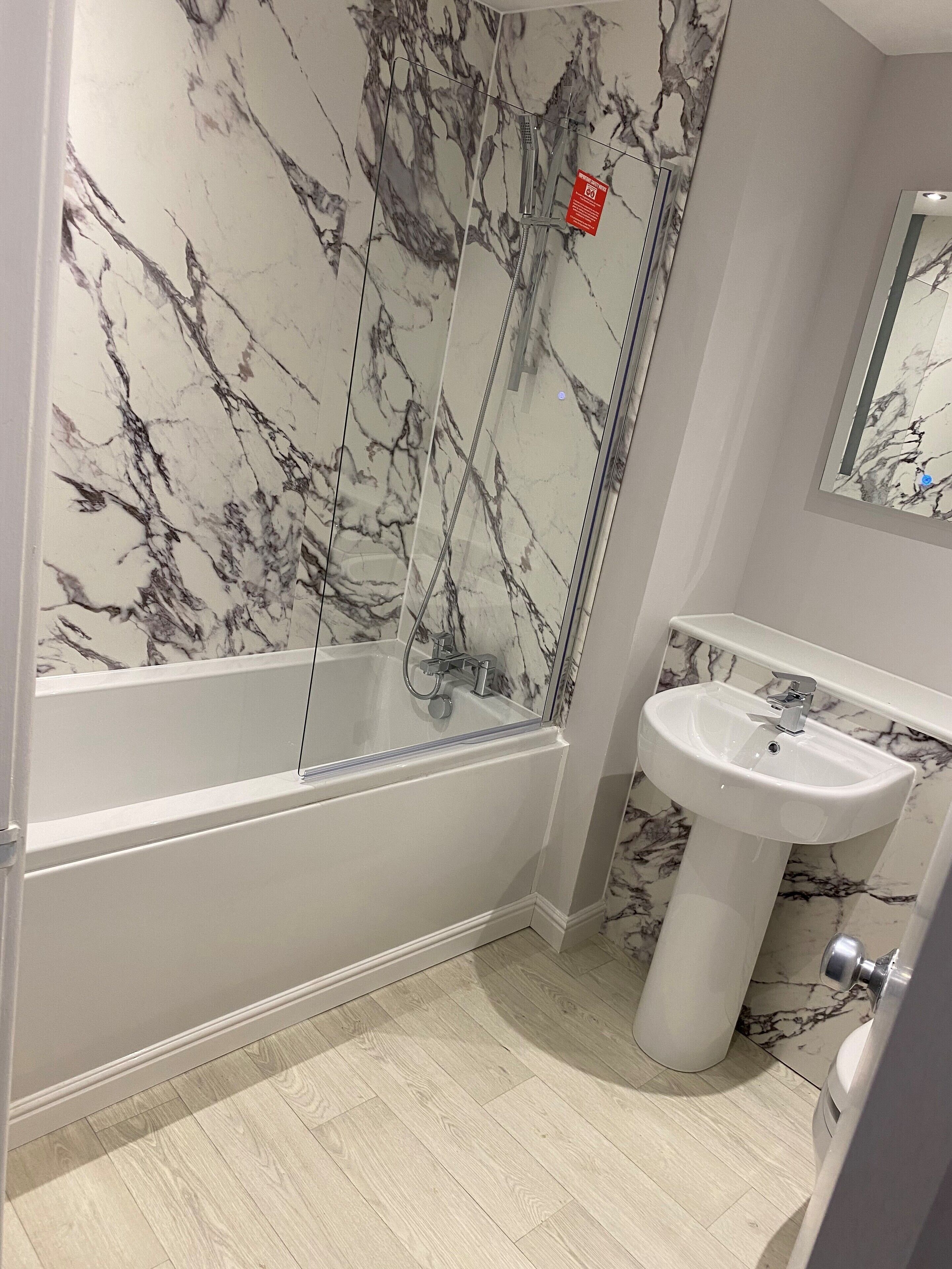 A bathroom with a marble shower wall, a white bathtub, and a pedestal sink.