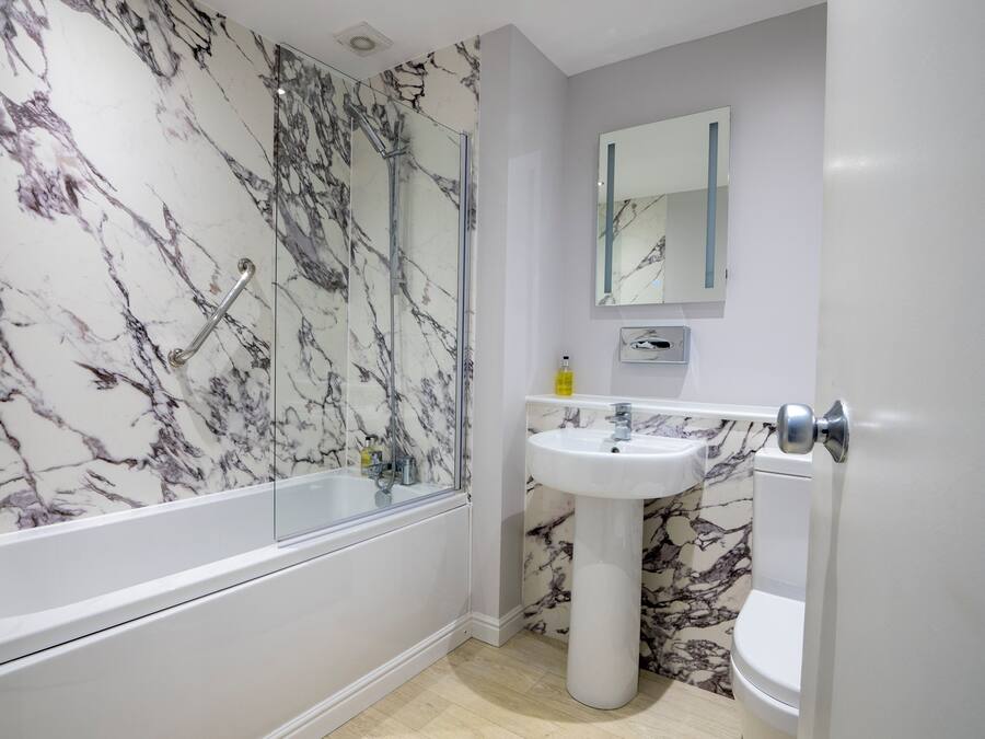 Un baño con pared de mármol, cabina de ducha de cristal, lavamanos pedestal y un inodoro.