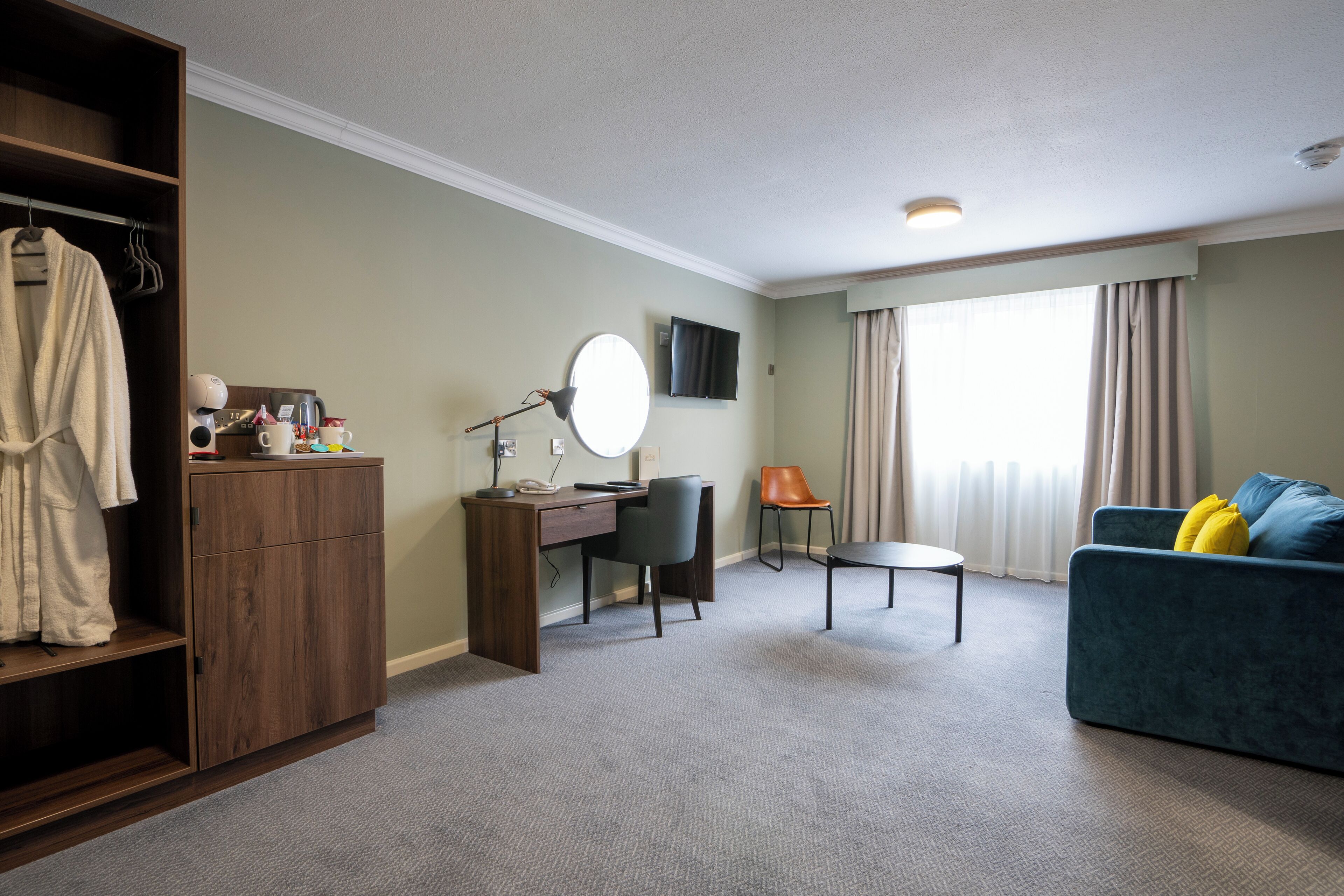 A hotel room with a wooden wardrobe, a desk with a chair, a round mirror, a television, a sofa with cushions, and a window with curtains.