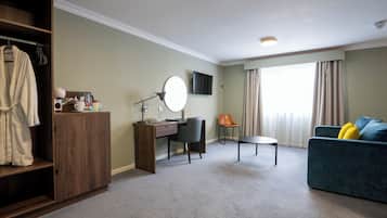 A hotel room with a wooden wardrobe, a desk with a chair, a round mirror, a television, a sofa with cushions, and a window with curtains.