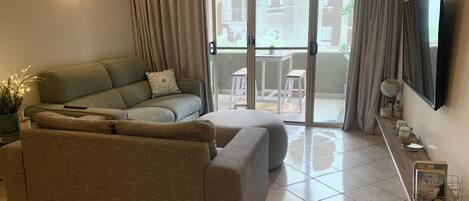Una sala de estar con un sofá modular, una mesa de centro, un televisor montado en la pared y un ventanal corredizo que da a un área exterior con una mesa y sillas.
