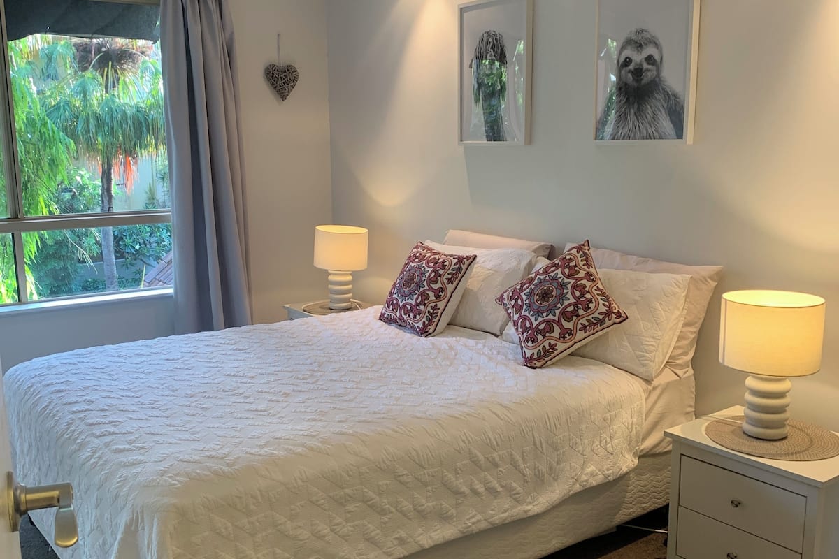 A bedroom with a large bed, two bedside lamps, a nightstand, a window with curtains, and a ceiling fan.