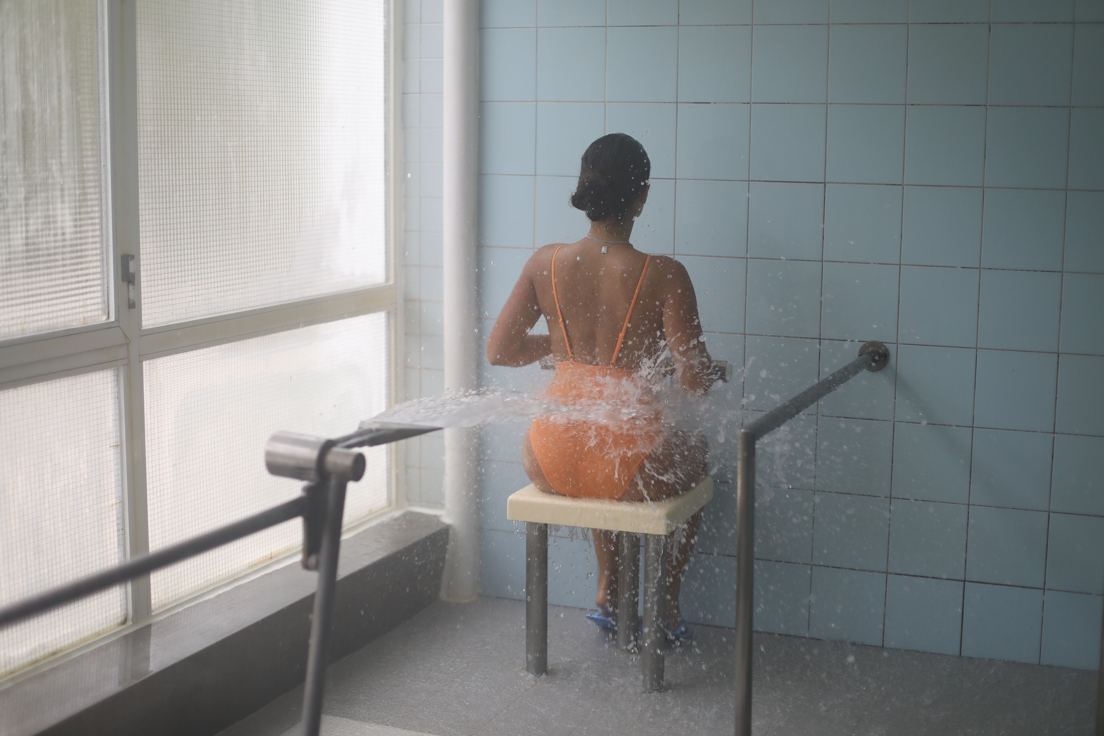 A person using a water fountain in a public space with tiled walls and a metal railing.