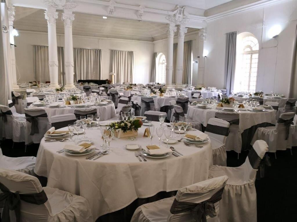 Un salón de banquetes con mesas redondas dispuestas para comer, cortinas blancas y columnas clásicas.