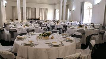 Un salón de banquetes con mesas redondas dispuestas para comer, cortinas blancas y columnas clásicas.