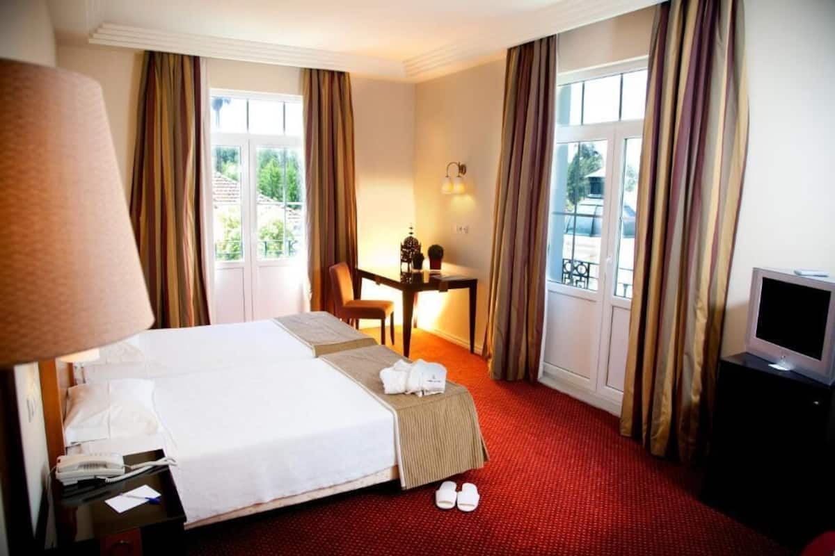 A hotel room with a large bed, a desk with a lamp, a television, and a view of the outdoors through the windows.