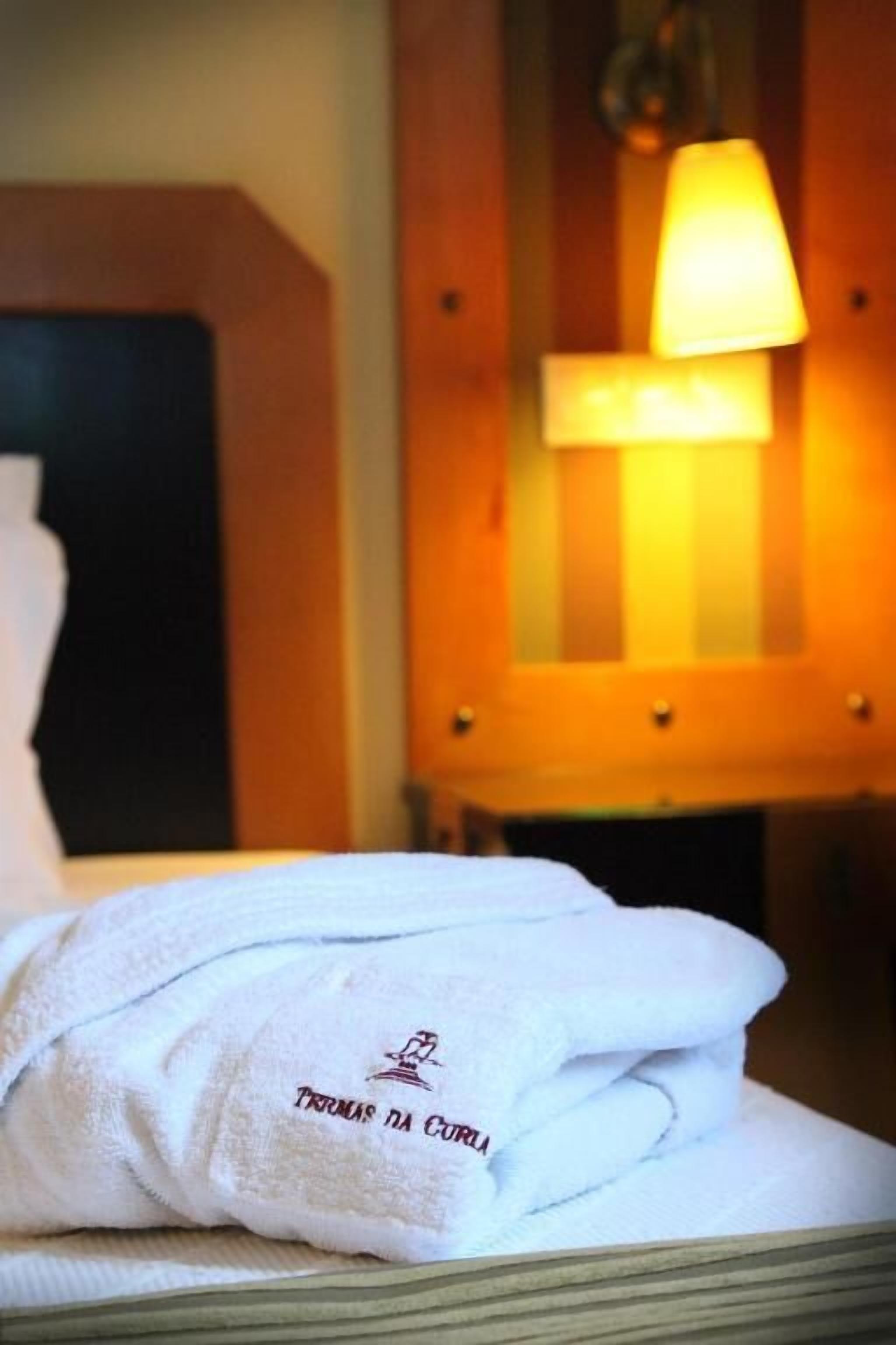 Una cama bien hecha con una toalla blanca que muestra el texto "Thomas da Corua", una mesita de noche de madera con una lámpara, y un cabecero de madera.
