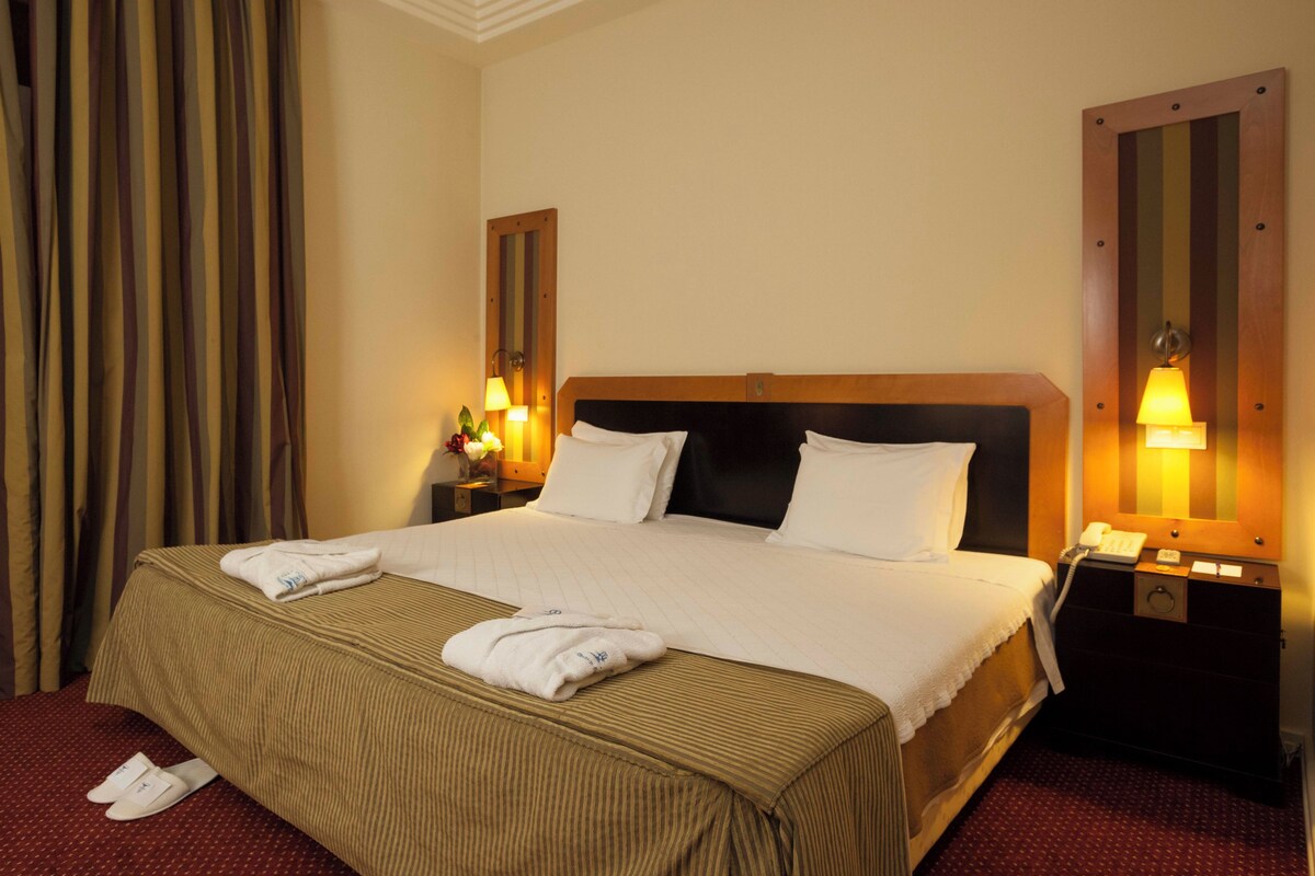 A hotel room with a large bed, two bedside lamps, a nightstand with a phone, and a mirror on the wall.