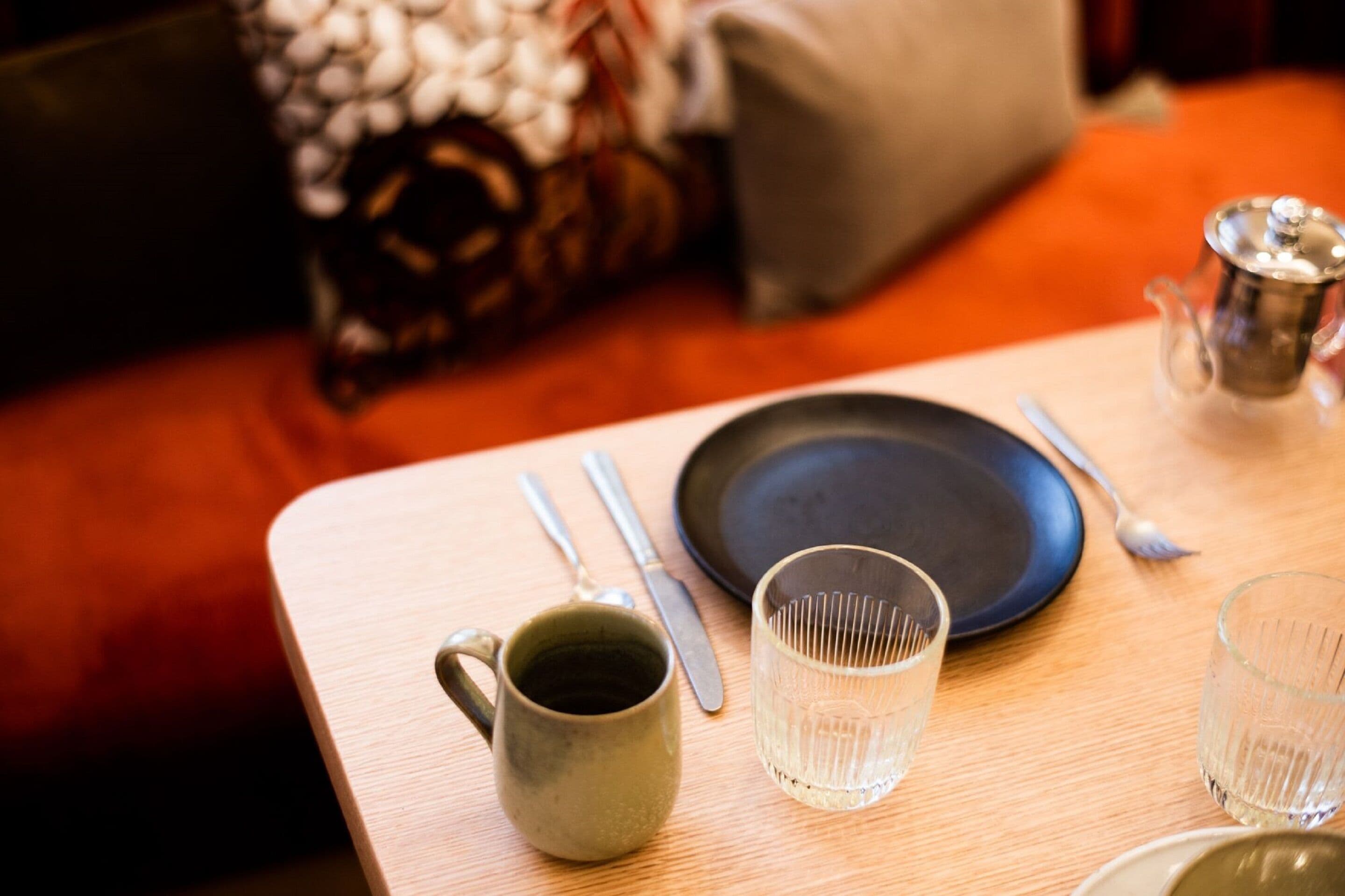 A table set with a dark plate, a clear glass, a metal pitcher, a mug, and cutlery.