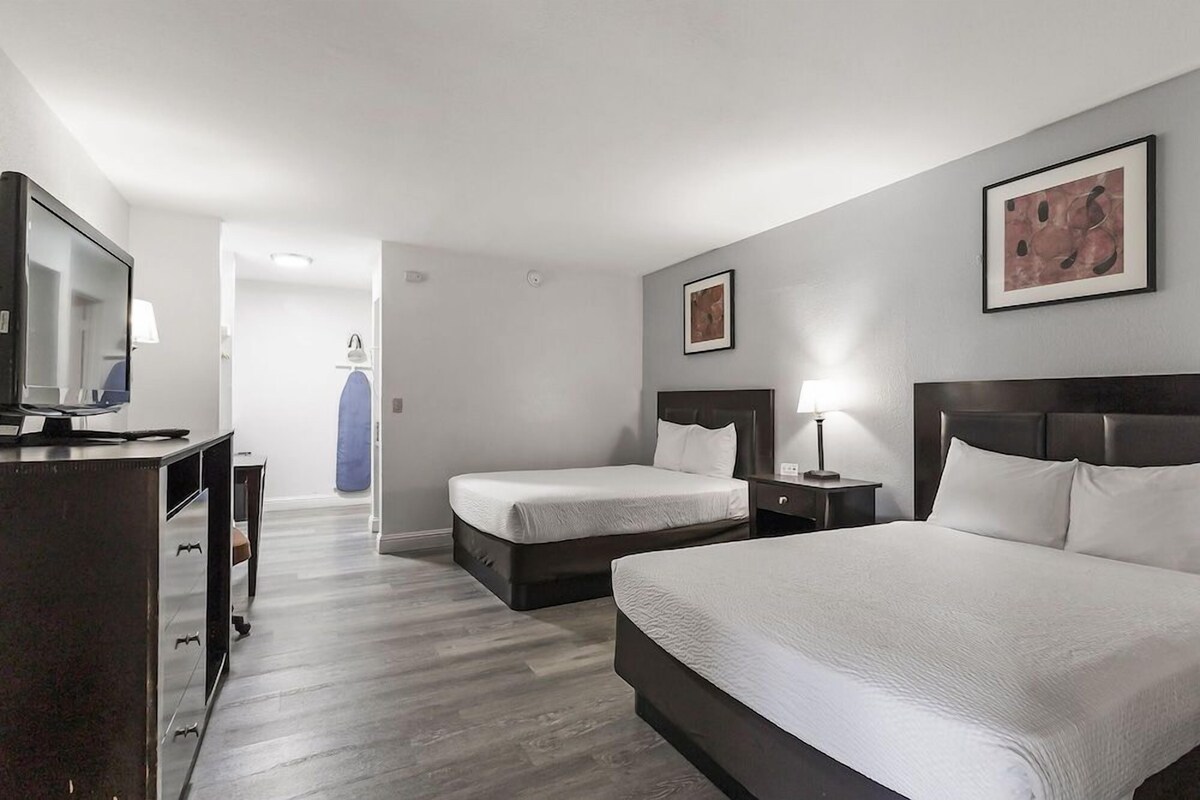 Quarto de hotel com duas camas, uma escrivaninha, televisão e obras de arte nas paredes.