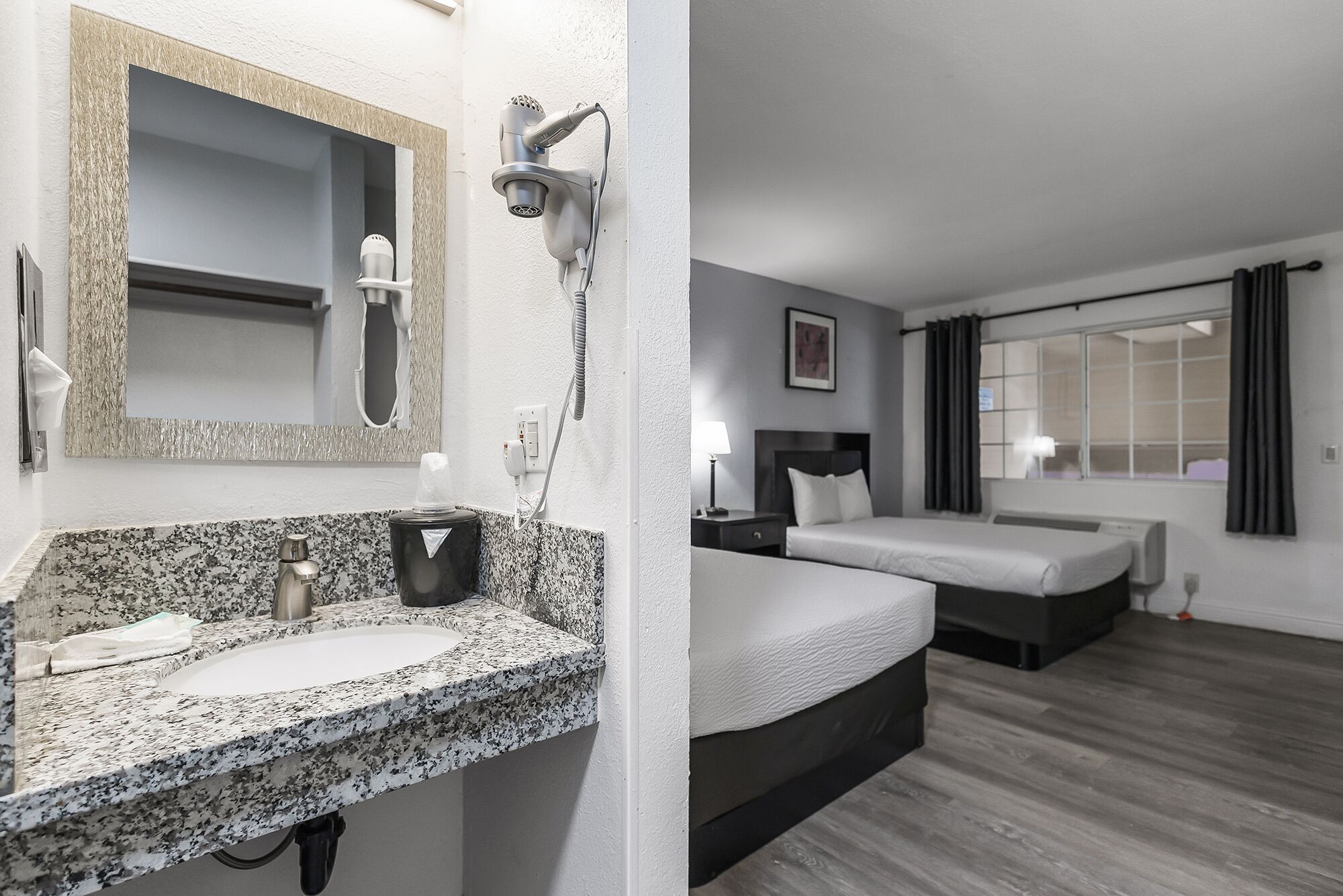 A hotel room with a granite countertop sink, a hairdryer, a mirror, a bed, a bedside table, and a window with curtains.