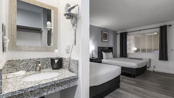 Habitación de hotel con lavamanos de encimera de granito, secador de pelo, espejo, cama, mesita de noche y ventana con cortinas.