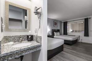 Habitación de hotel con lavamanos de encimera de granito, secador de pelo, espejo, cama, mesita de noche y ventana con cortinas.