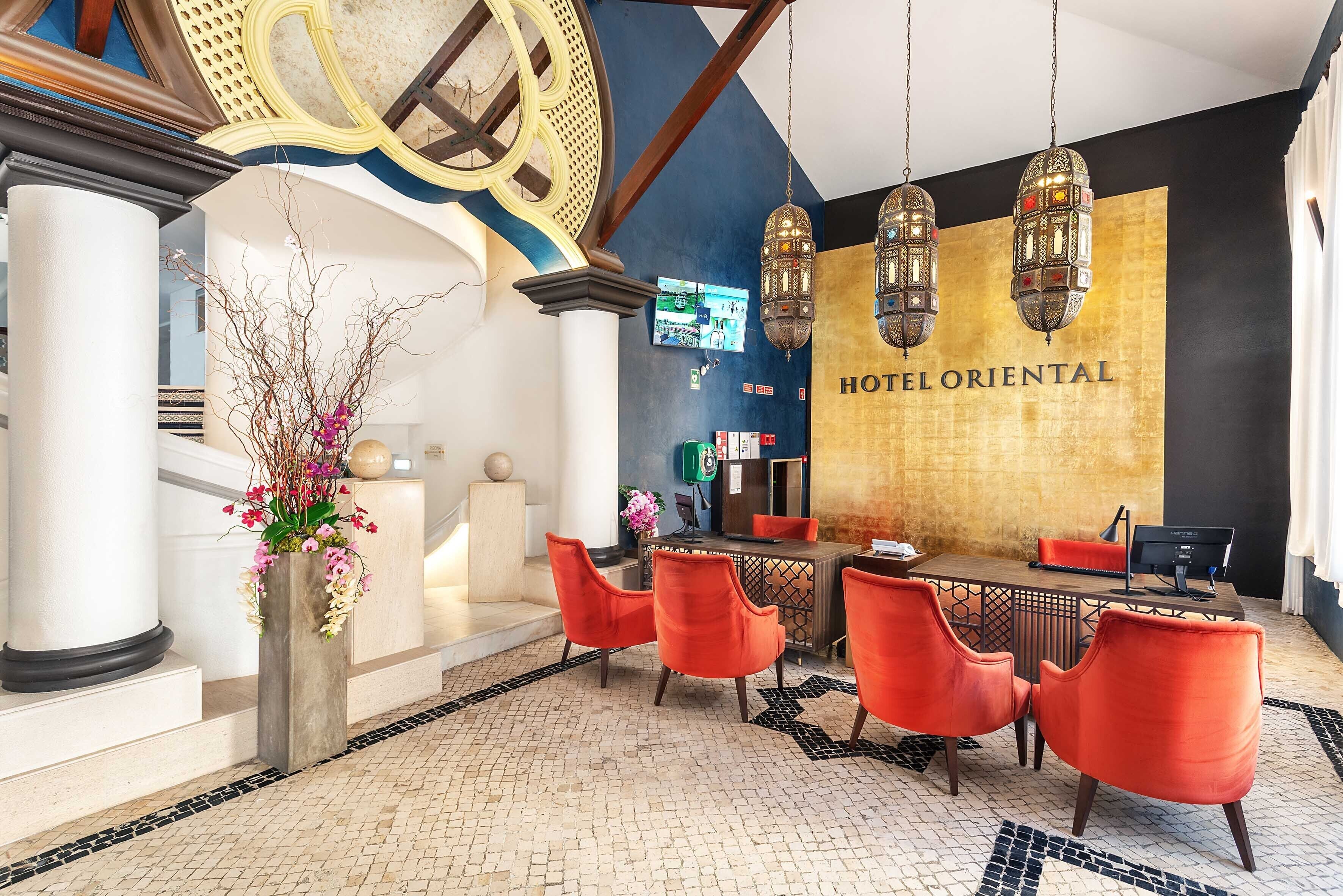 Un vestíbulo de hotel con una recepción, sillas rojas, una pared decorativa grande y un suelo de baldosas.