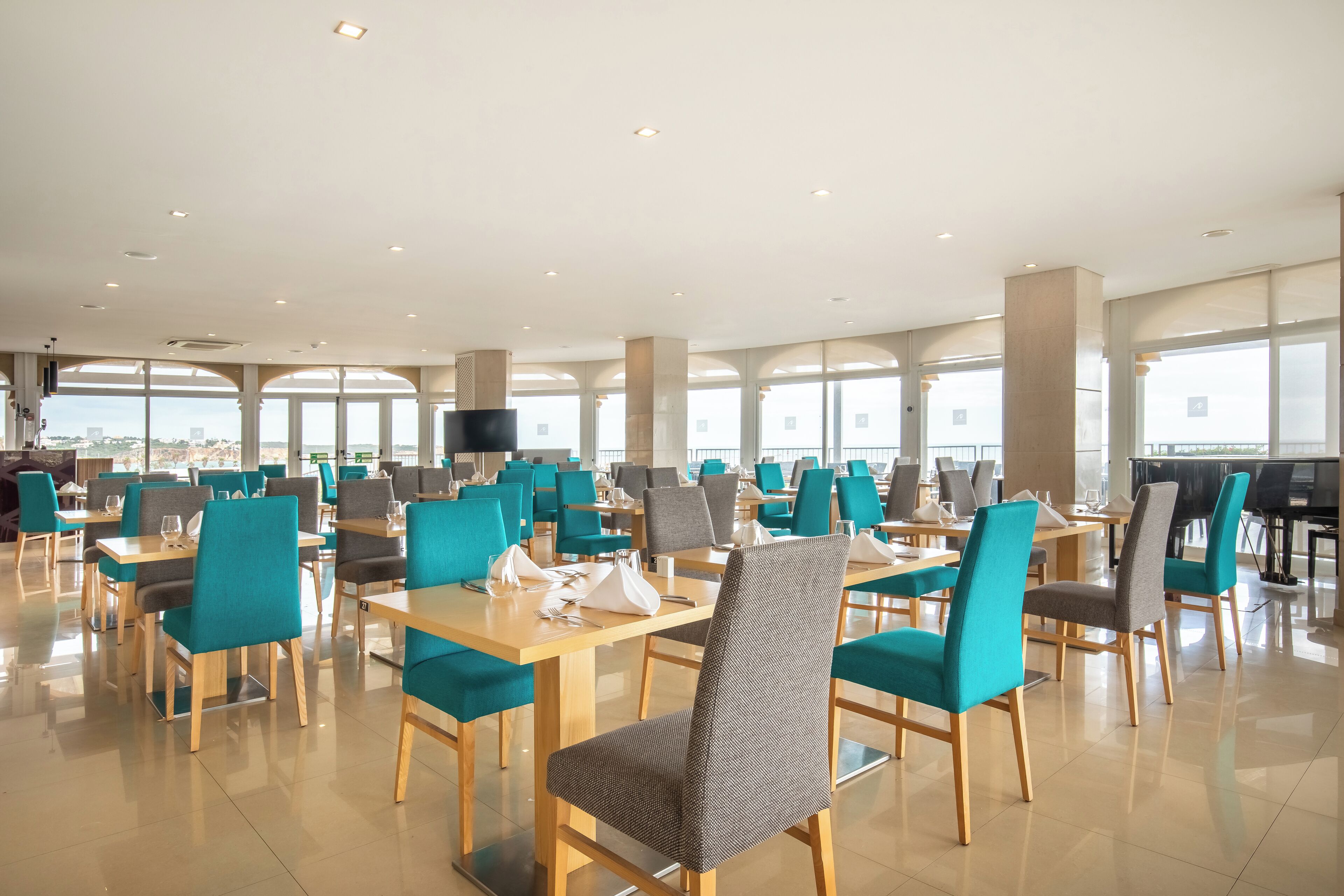 A spacious dining area with multiple tables and chairs, large windows, and a piano.