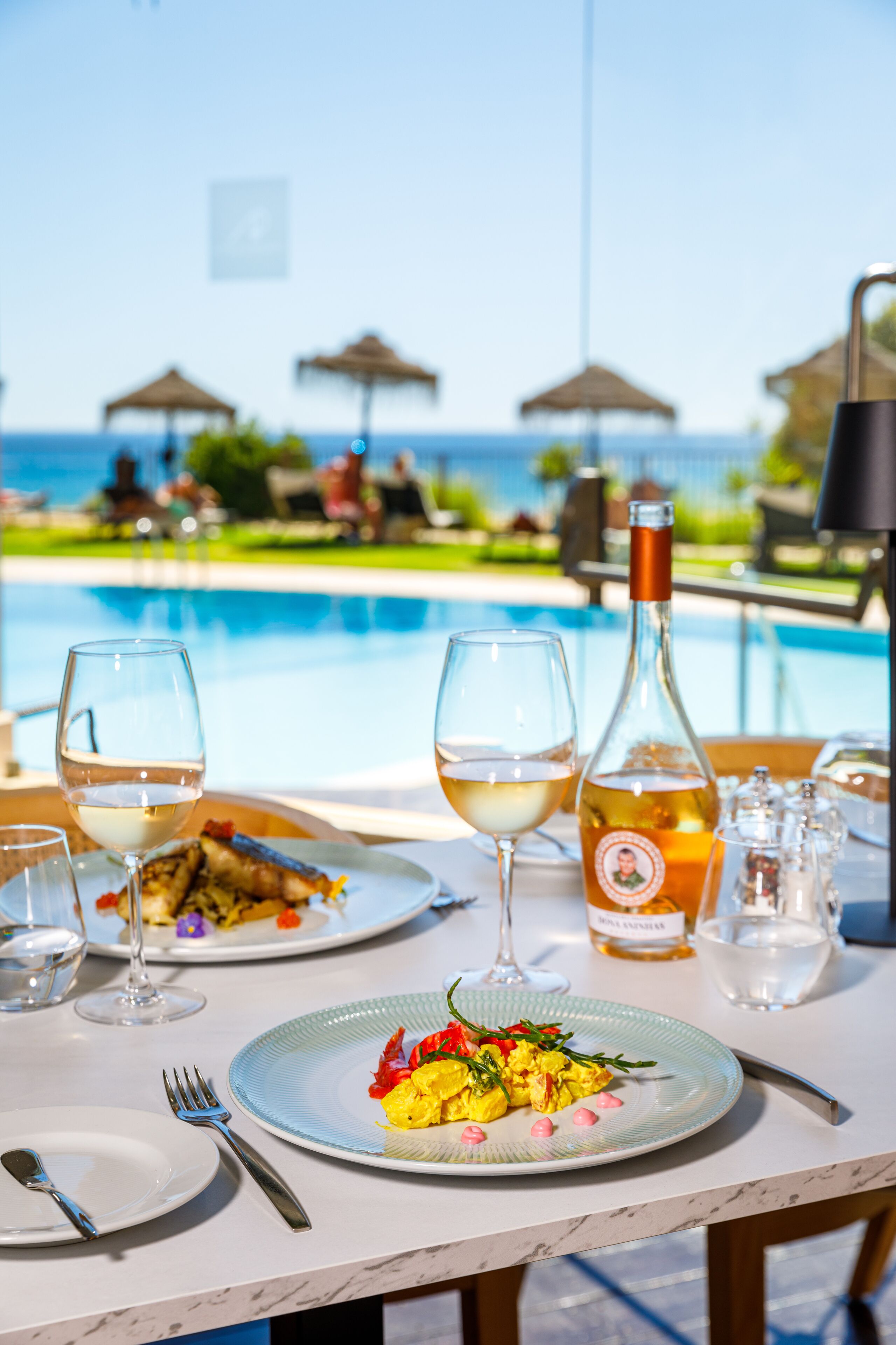 A dining table with plates, glasses, and a bottle of wine, overlooking a pool and lounge area.