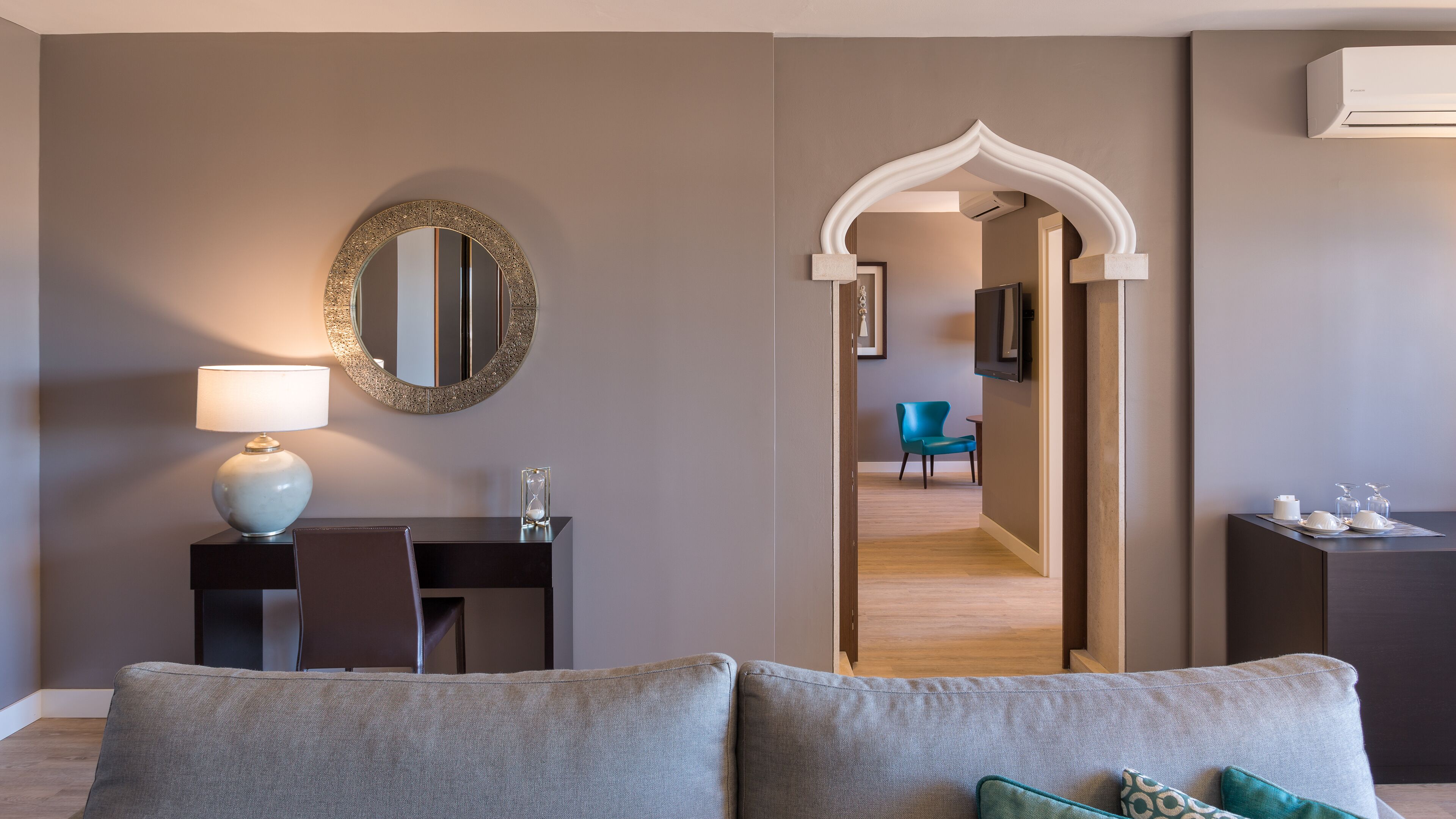 A modern hotel room with a sofa, a desk with a lamp, a mirror, and a TV.