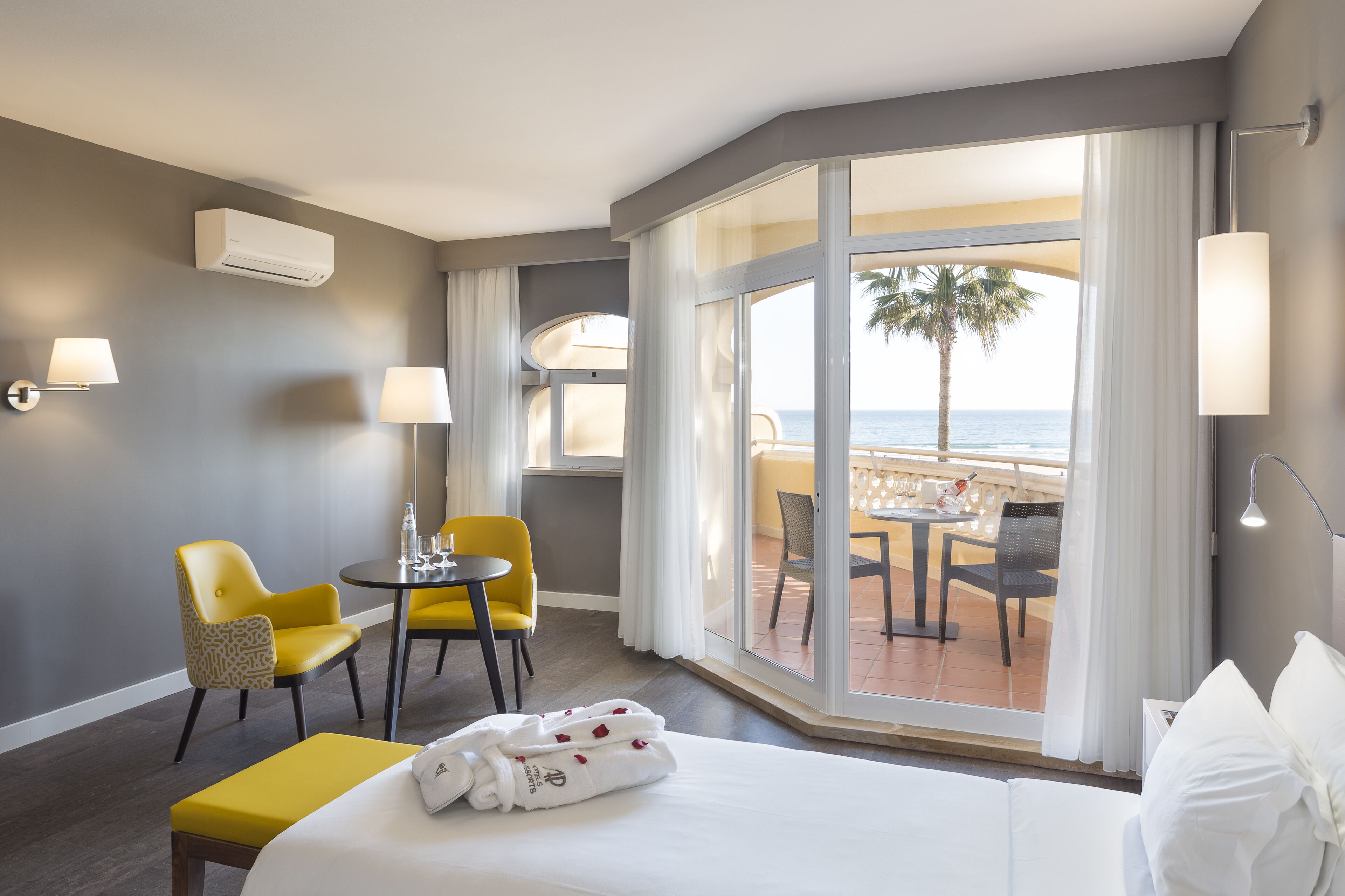 A hotel room with a bed, a yellow chair, a table, and a view of the beach.