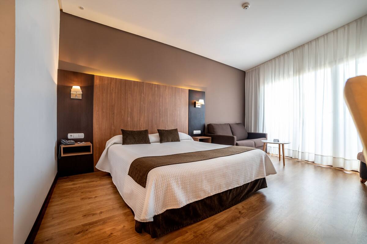 A modern hotel room with a large bed, a seating area with a chair, a small table, and a large window with curtains.
