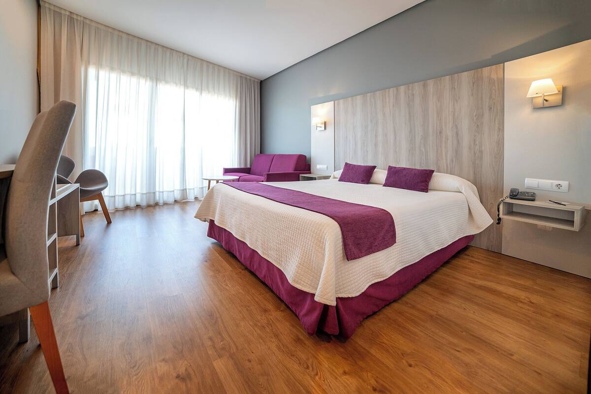 A modern hotel room with a large bed, a desk, a chair, and a sofa.