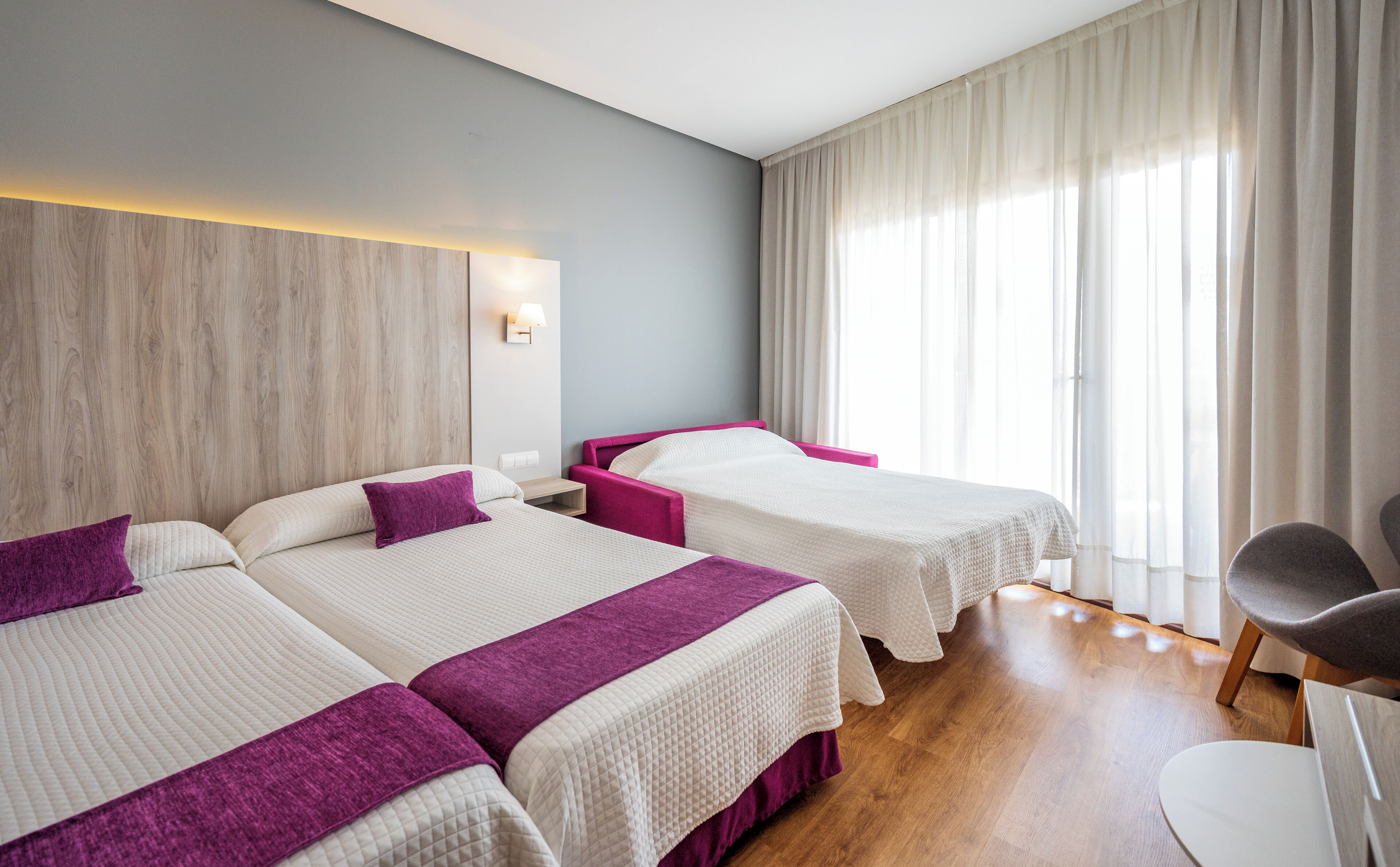 A hotel room with two beds, a wooden headboard, a bedside table with a lamp, and a chair.