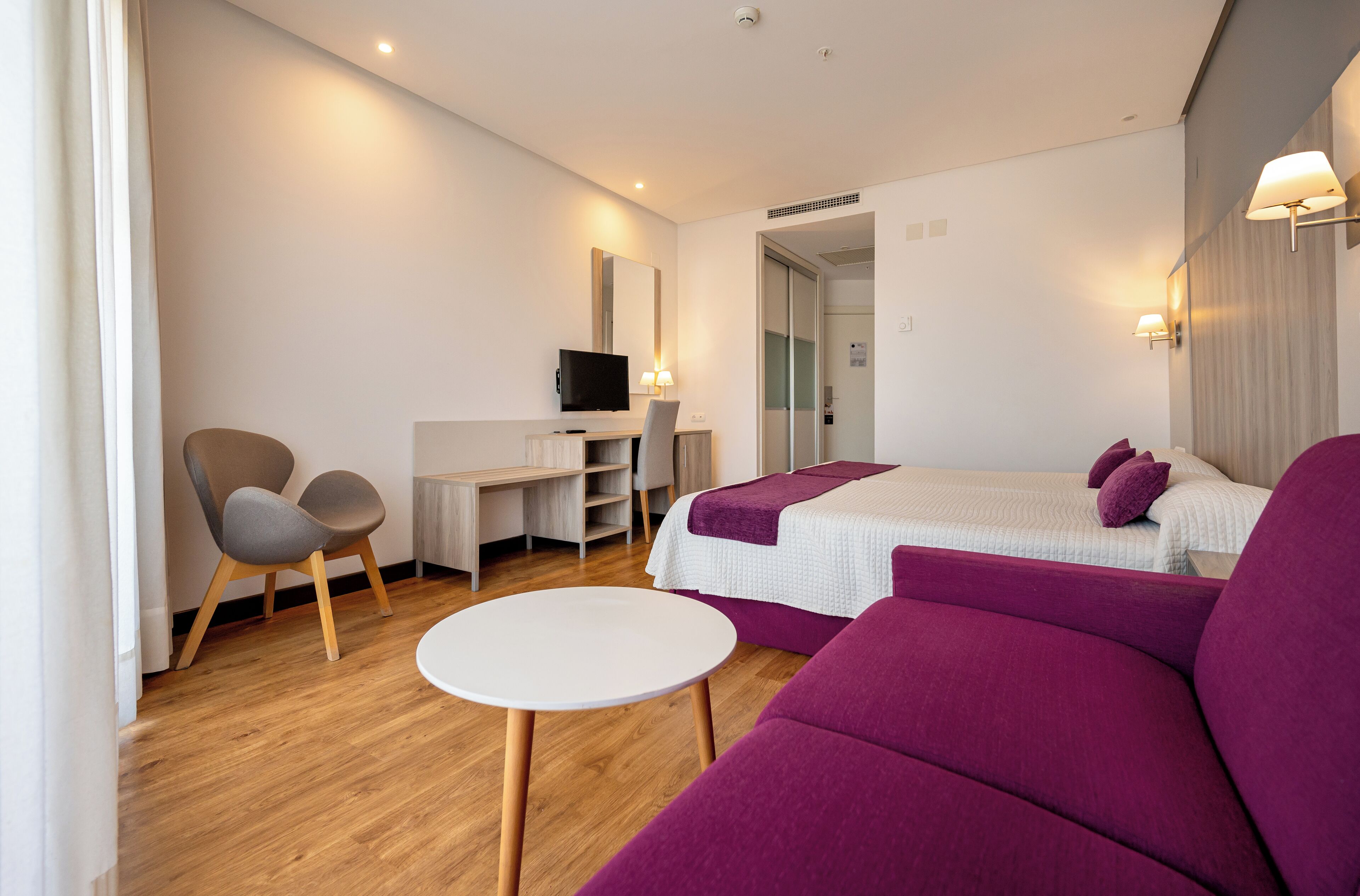 A modern hotel room with a wooden floor, a white desk with a flat-screen TV, a purple sofa, and a bed with white bedding.