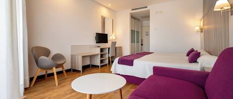 A modern hotel room with a wooden floor, a white desk with a flat-screen TV, a purple sofa, and a bed with white bedding.
