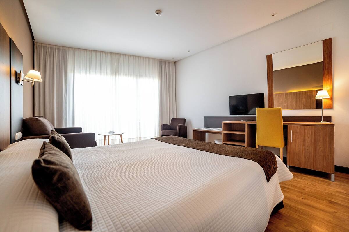 A hotel room with a large bed, a desk with a computer, a TV, and a seating area with chairs.